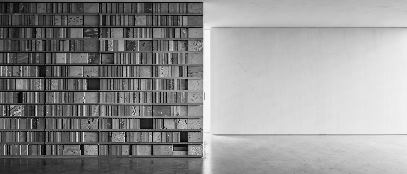 Conceptual editorial illustration — a luminous threshold between a dense library-like wall of ordered stone forms and an open empty plane. Monochrome study of the frontier between accumulated knowledge and the open machine economy.
